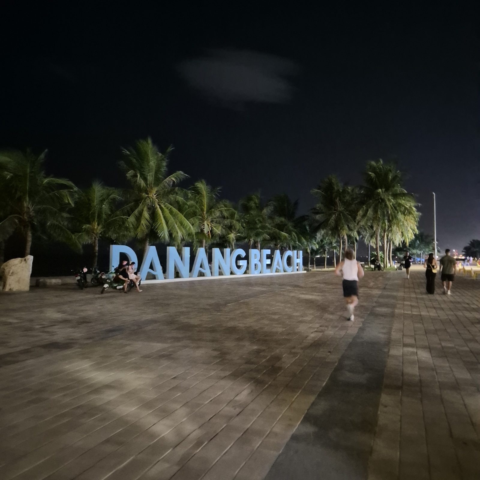 밤의 미케 산책로. 파란 DANANG BEACH 블록 레터와 야자수, 멀리 흐린 보행자들만 보이는 해변 랜드마크